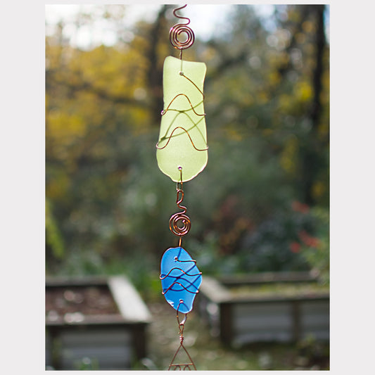 Decorative glass hanging with green and blue pieces against a blurred natural background