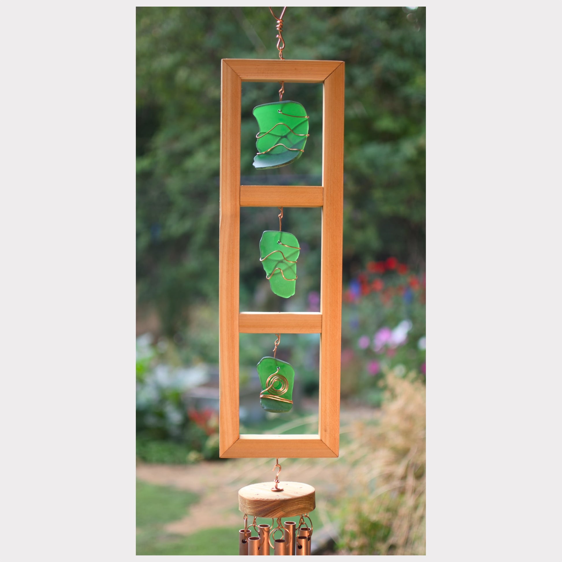 Decorative wind chime with green glass leaves and wooden frame against a garden background