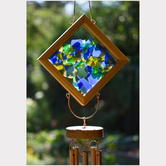 Diamond-shaped wind chime with a wooden frame and colorful blue, green, and yellow mosaic glass panels, hanging outdoors against a blurred green background.