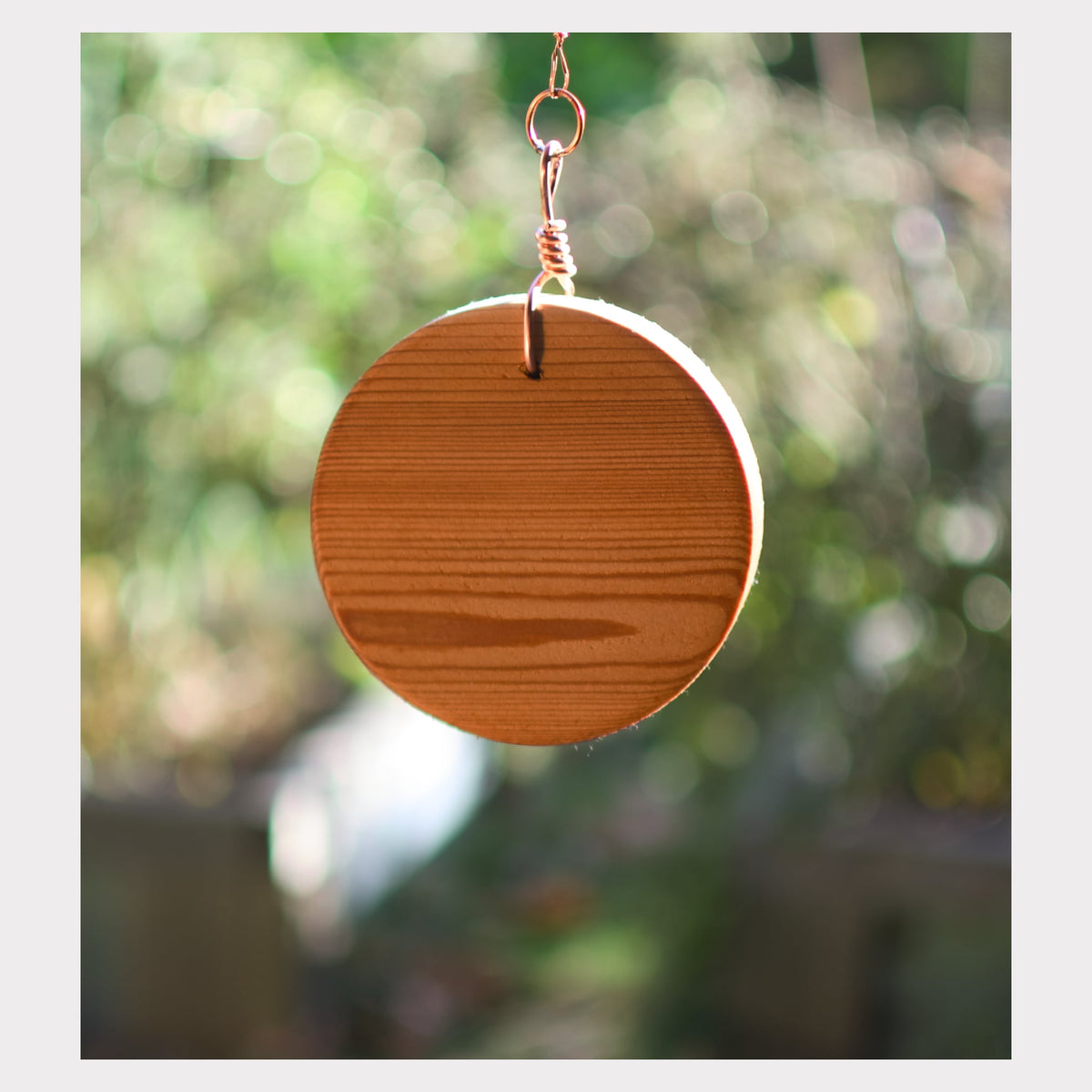 A round wooden wind chime hanging from a copper wire loop against a blurred green and brown outdoor background.