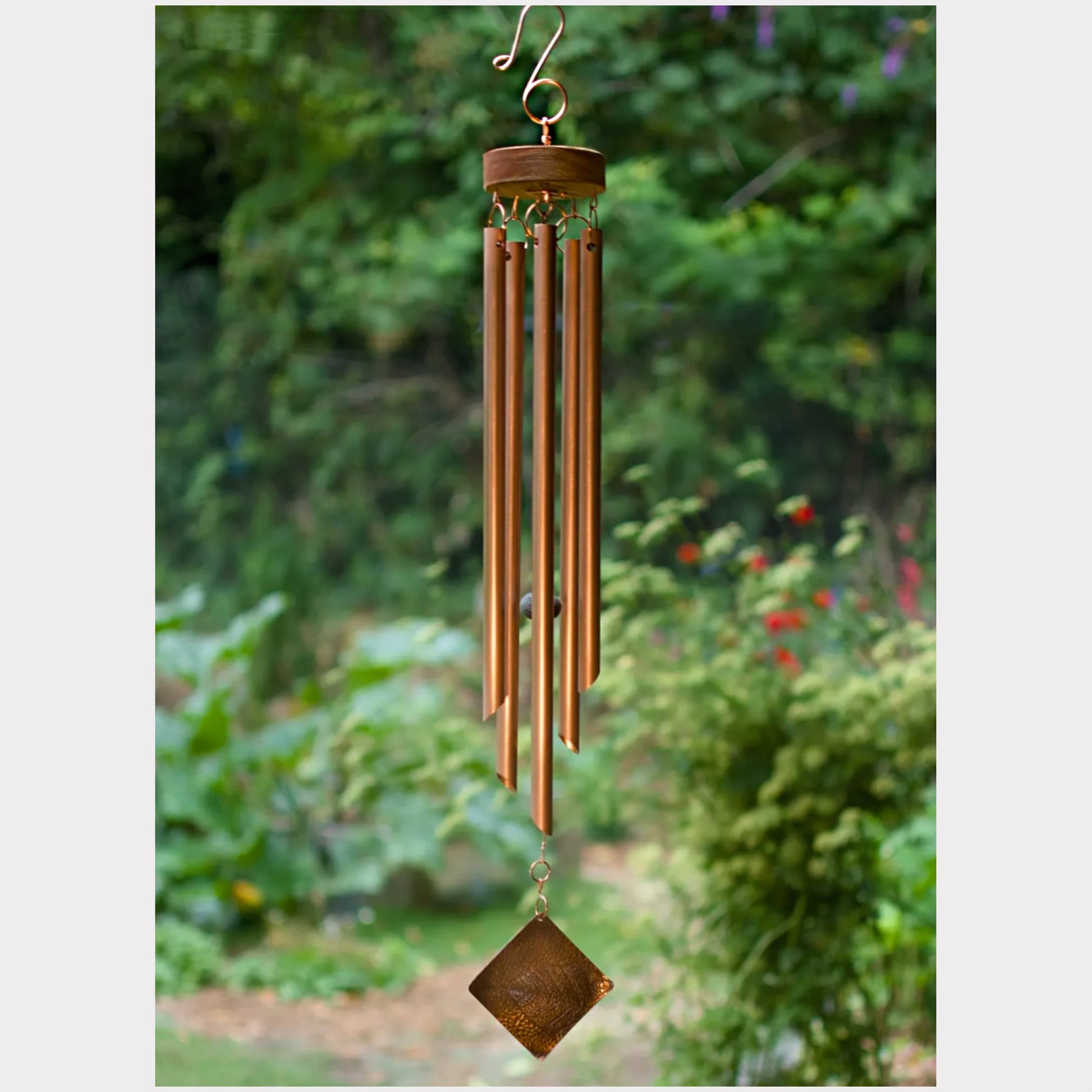 A hanging wind chime with multiple metallic rods and a diamond-shaped pendant, photographed outdoors against a blurred green garden background.