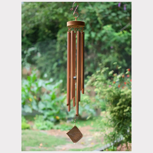 A copper wind chime with multiple hanging tubes and a diamond-shaped crystal striker, shown outdoors against a blurred garden background.