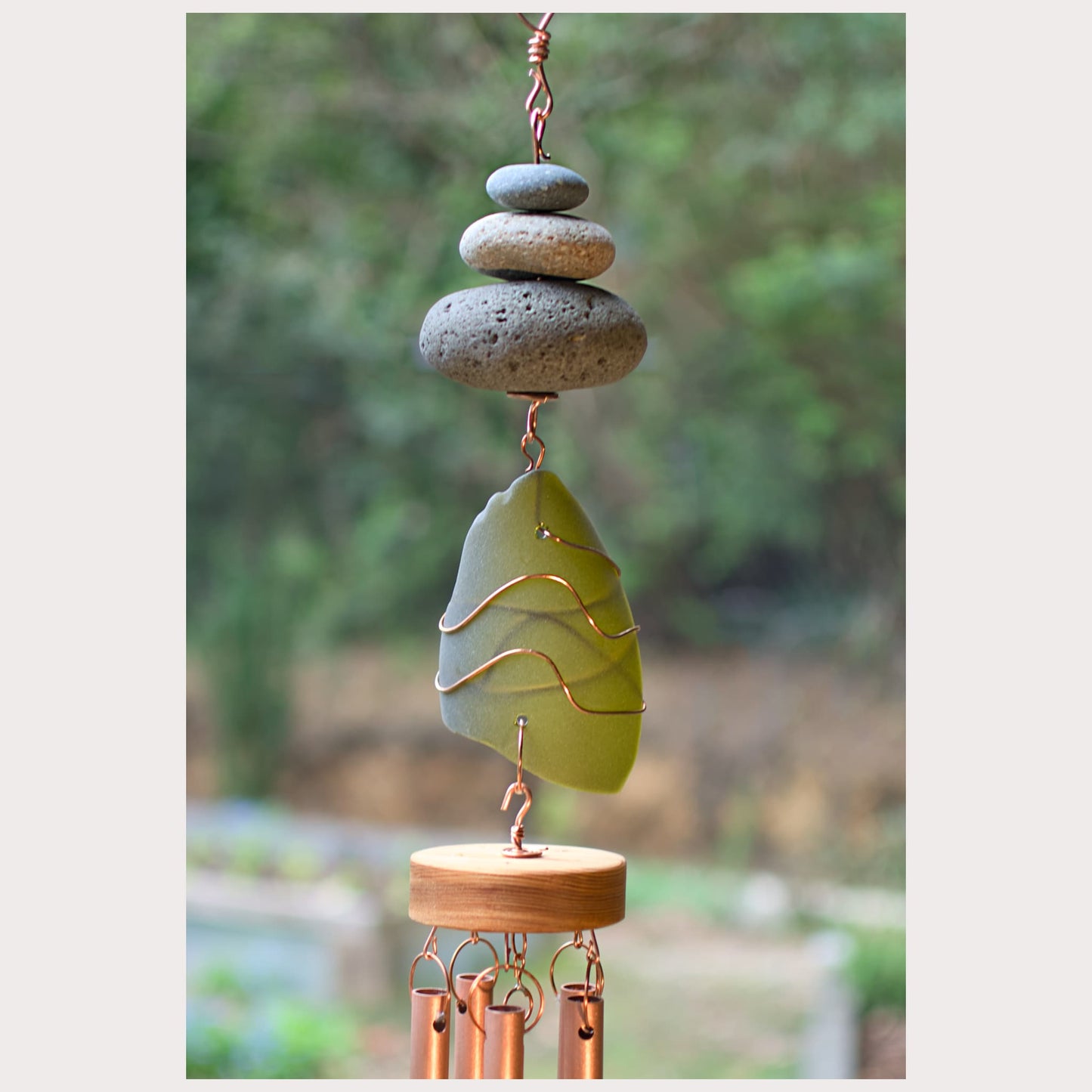 Decorative wind chime with stacked stones and a green leaf-shaped ornament against a blurred natural background.