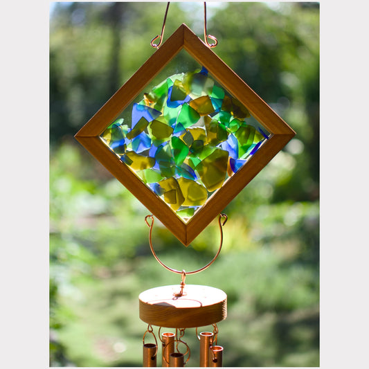 A diamond-shaped wooden suncatcher with colorful green, blue, and yellow stained glass panels, hanging from a copper wire loop above a wind chime with copper tubes, set against a blurred green outdoor background.