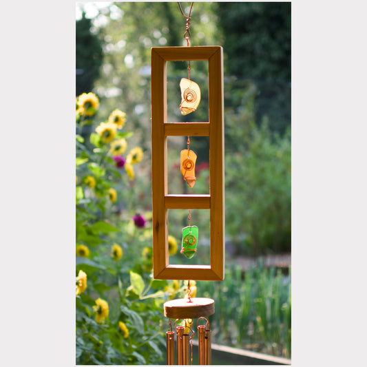 A wooden hanging wind chime with three rectangular panels, decorated with natural shell and paper weights, suspended outdoors in front of a blurred garden background with sunflowers.