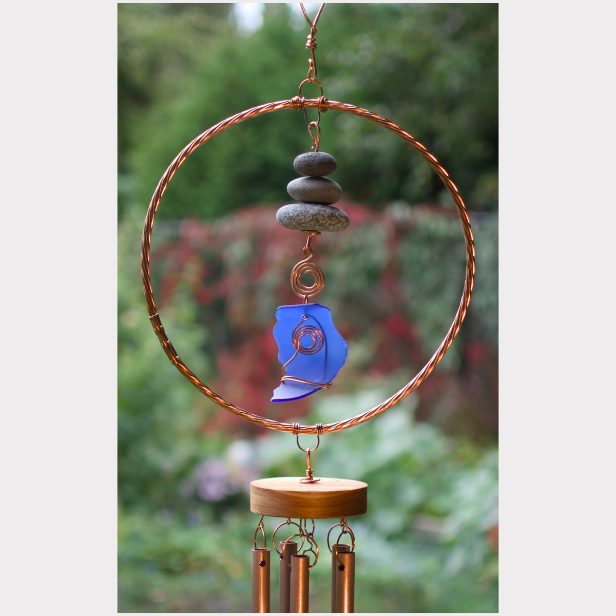 A handmade copper wind chime featuring a large circular hoop, stacked gray stones, a blue sea glass piece, and three hanging copper tubes, photographed against a blurred green garden background.