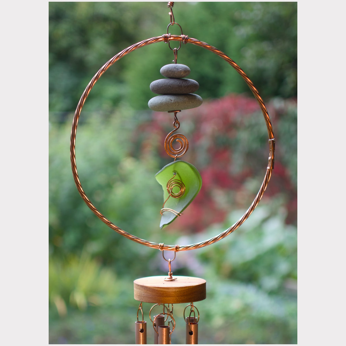 A handmade wind chime hanging outdoors against a blurred green garden background. The chime features a large copper wire hoop, a stack of gray stones, a green glass piece, and a round wooden bottom plate with hanging copper tubes.