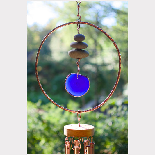 A copper wind chime with a large circular hoop, stacked stones, a vibrant blue glass disc, and copper wind chime tubes, set against a blurred green garden background.
