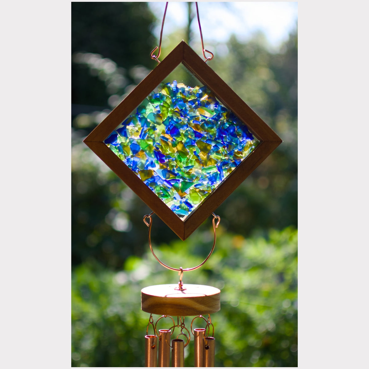 A diamond-shaped wind chime with a wooden frame and a colorful mosaic glass center, hanging outdoors with copper-colored wind chime tubes below against a blurred green background.