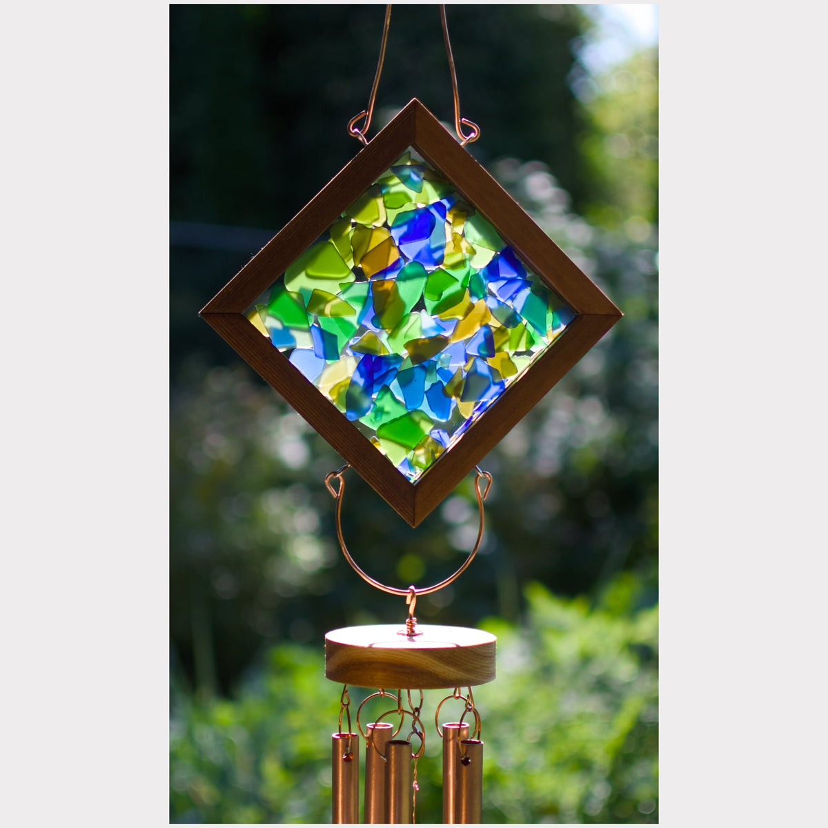 A diamond-shaped wind chime with a wooden frame and colorful stained-glass style panels in blue, green, and yellow, hanging outdoors against a blurred green garden background.