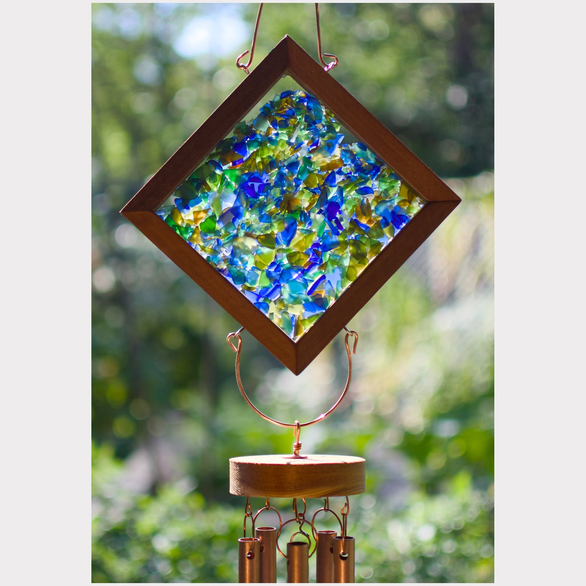 A diamond-shaped wind chime with a dark brown wooden frame and a colorful mosaic of blue, green, and yellow glass pieces, hanging outdoors against a blurred green garden background.