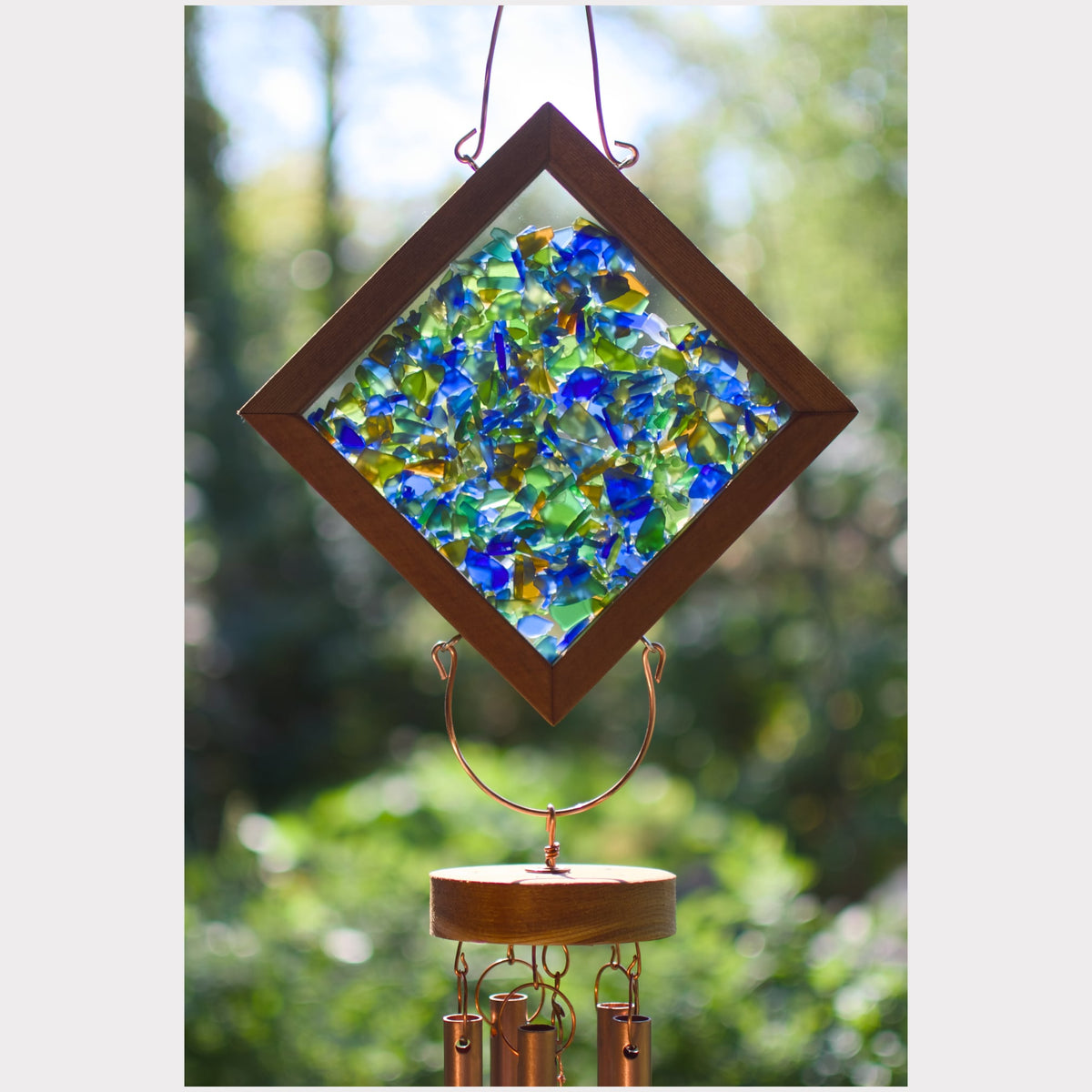 A diamond-shaped glass wind chime hanging outdoors against a blurred green foliage background. The chime features a brown wooden frame filled with blue, green, and yellow broken glass, suspended from a copper-colored metal wire frame with cylindrical wind chimes below.