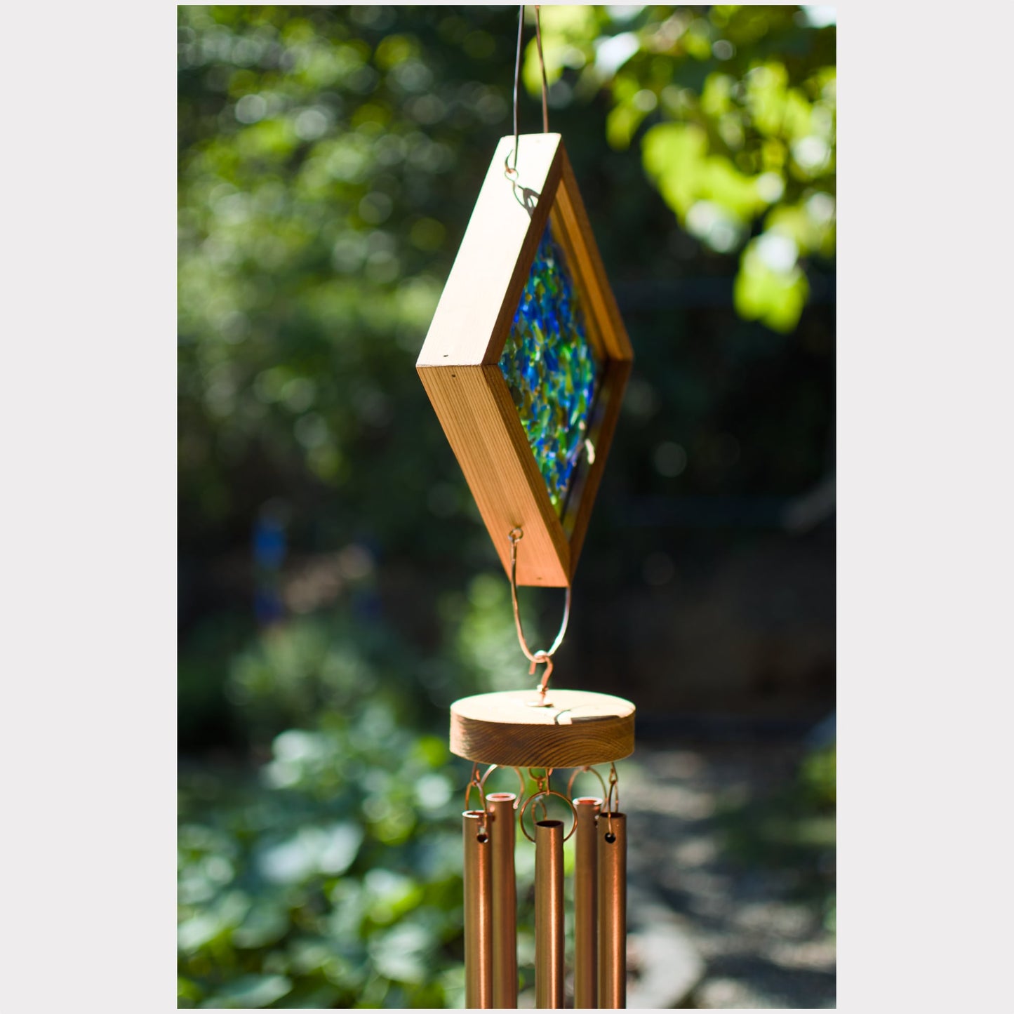 A diamond-shaped wind chime hanging outdoors, featuring a natural wood frame with a colorful mosaic center and copper-colored metal tubes below, set against a blurred green garden background.