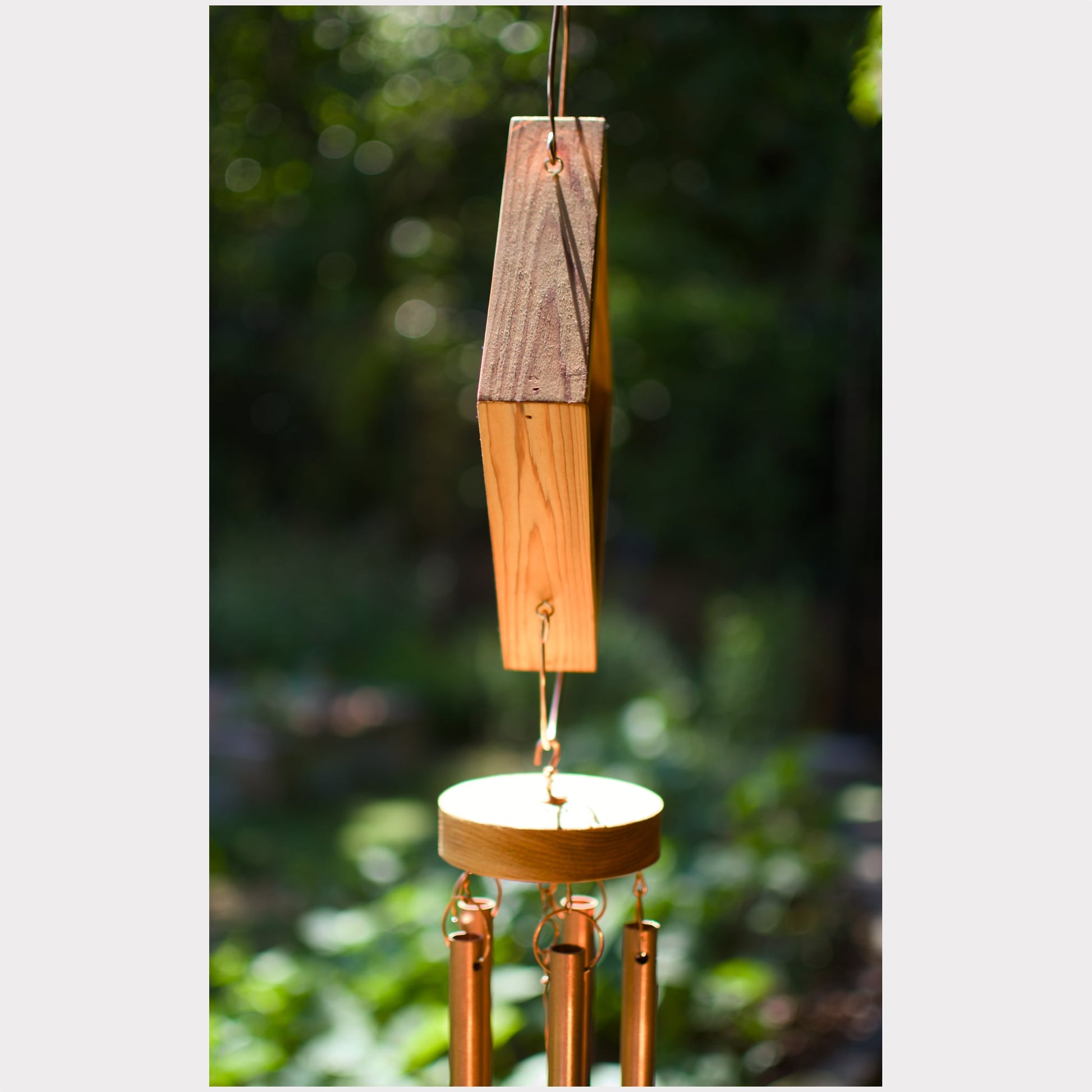 A wooden wind chime hanging outdoors against a blurred green forest background. The chime features a rectangular wooden striker with a pointed top, a round wooden striker disc, and several copper-colored metal tubes hanging below.