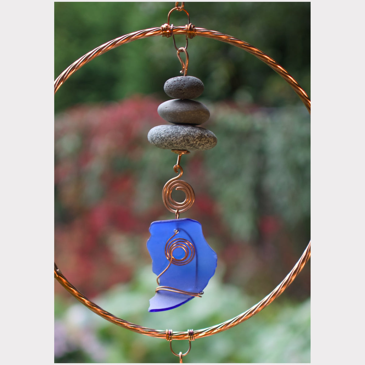 A handmade copper wire wind spinner featuring a large circular hoop, stacked gray stones, and a blue glass piece wrapped with copper wire, set against a blurred garden background.