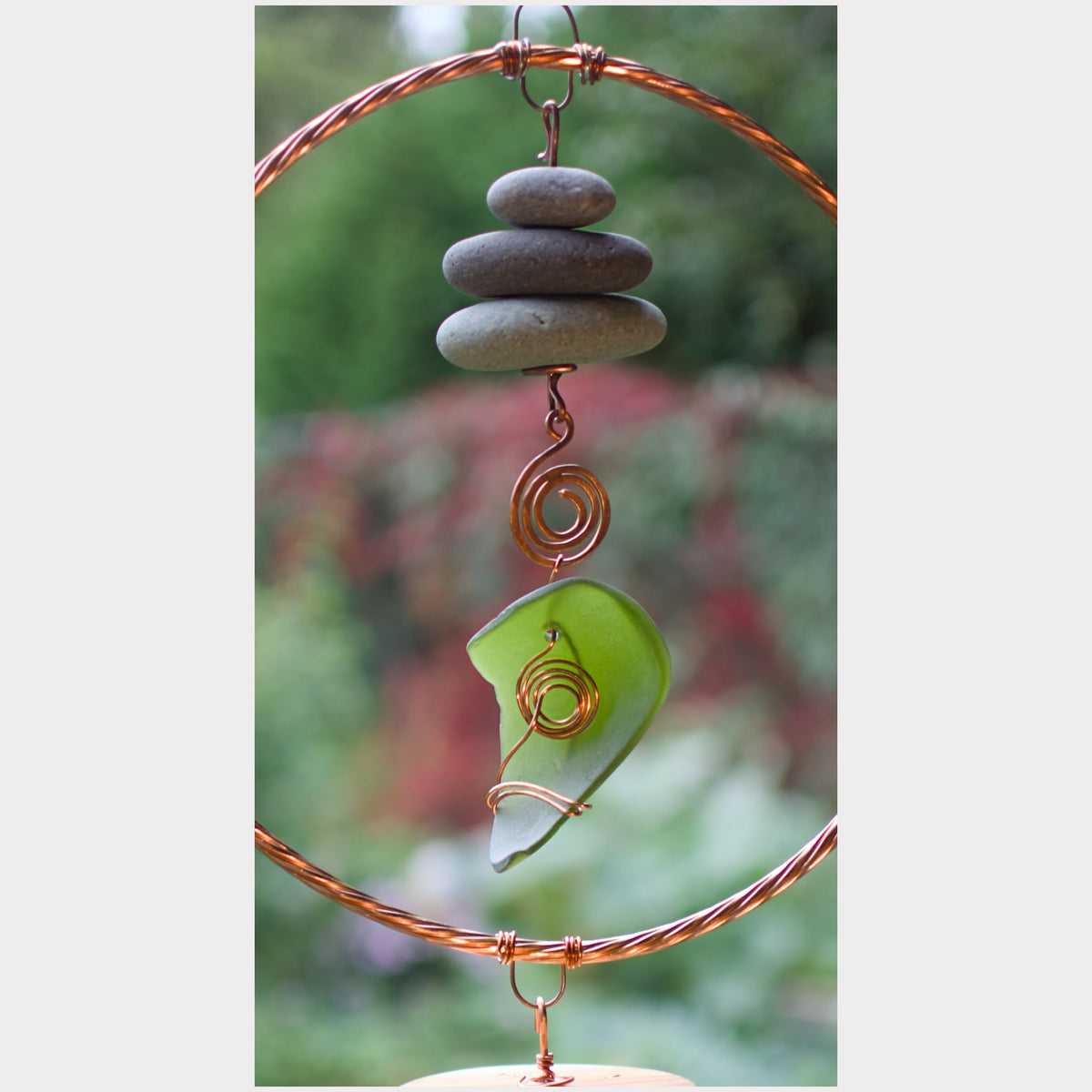 A handmade wind spinner ornament featuring a copper wire double-ring frame, three stacked gray stones, a green sea glass piece, and copper wire swirls, set against a blurred garden background.