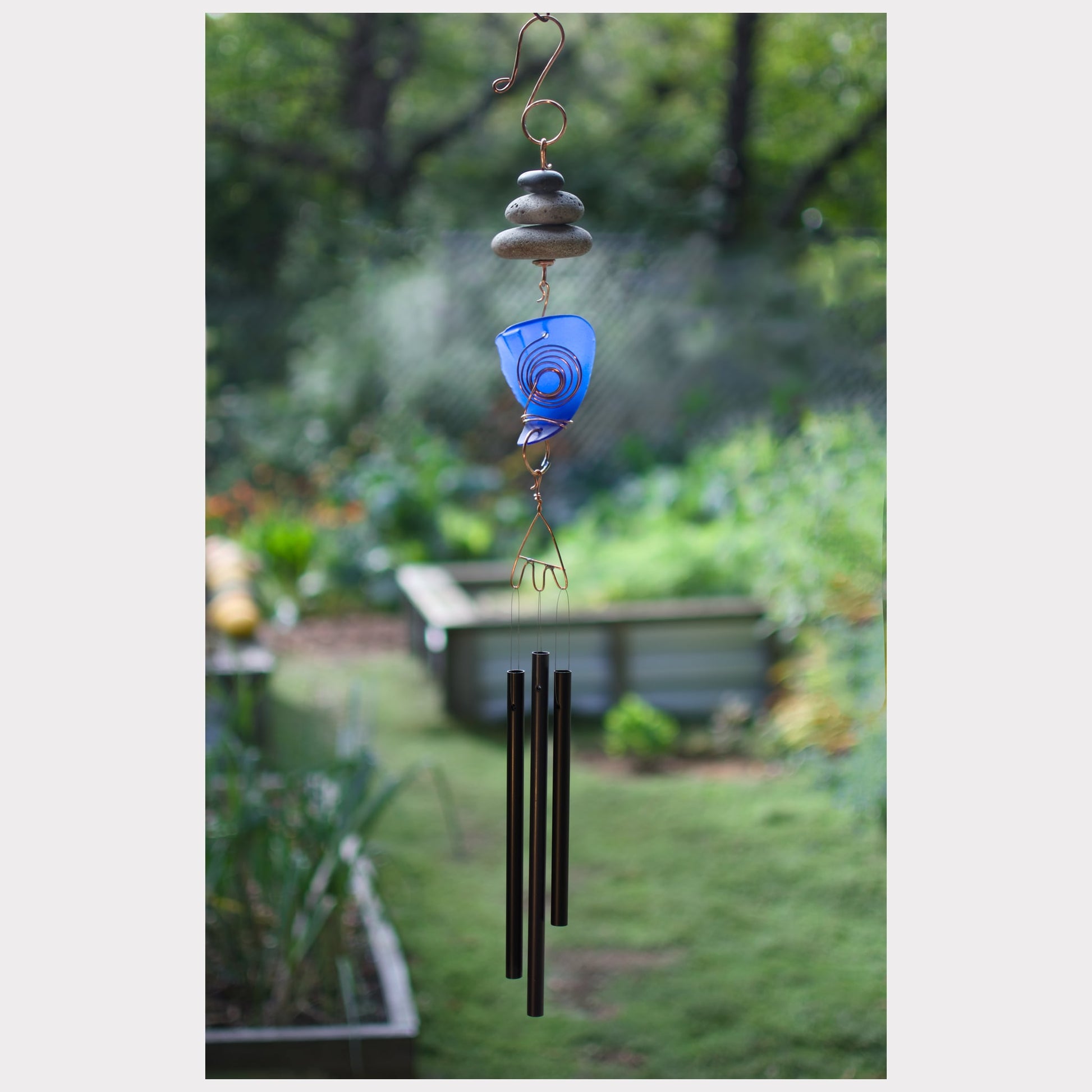 Decorative wind chime with blue glass element and black metal tubes against a garden background