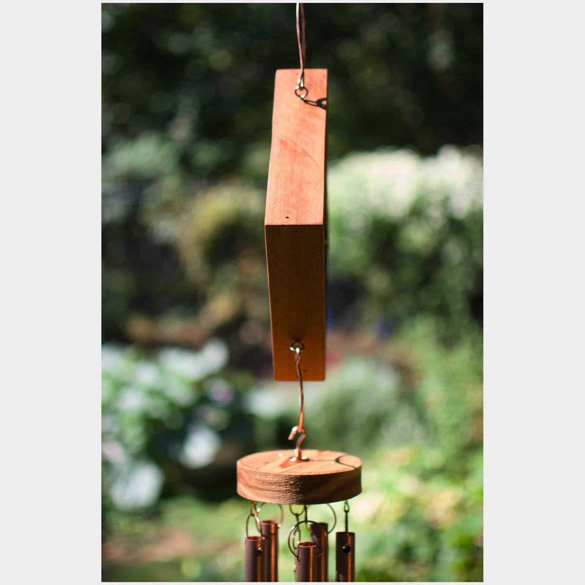 A handmade wooden wind chime hanging outdoors against a blurred green garden background. The chime features a rectangular wooden striker block suspended from a round wooden hoop, with several dark brown metal tubes hanging below.