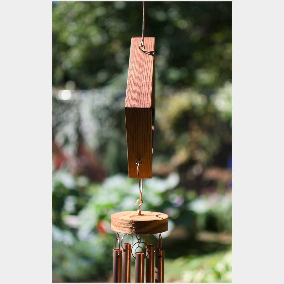 A wooden wind chime hanging outdoors, with a rectangular wooden striker at the top and several metallic tubes below, set against a blurred green garden background.