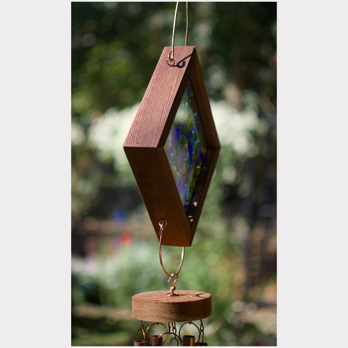 A diamond-shaped wooden wind chime with a colorful stained glass insert, hanging outdoors against a blurred green garden background.