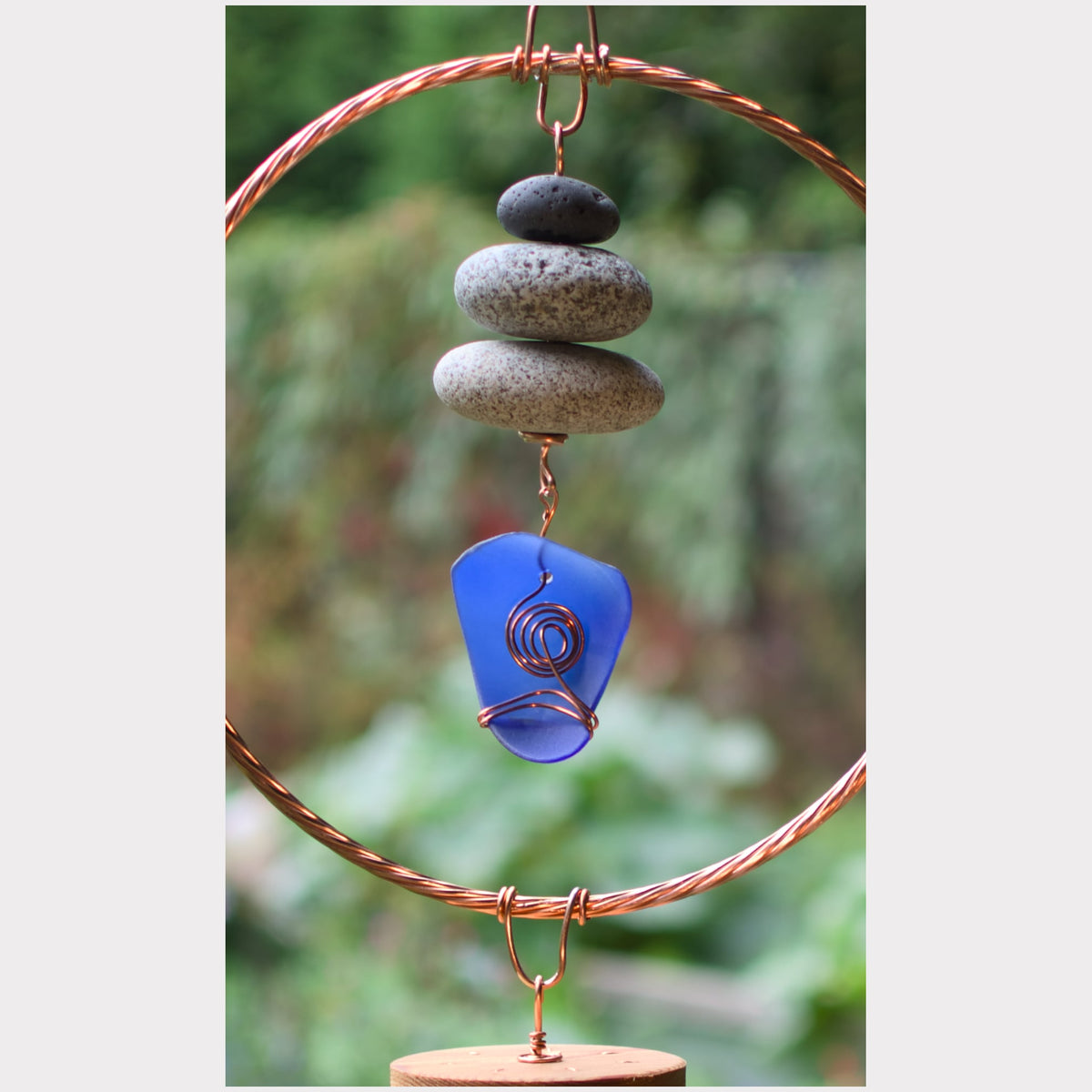 A handmade copper wire wind spinner featuring a stacked arrangement of gray and black river stones and a blue glass guitar pick with a copper spiral detail, set against a blurred green garden background.