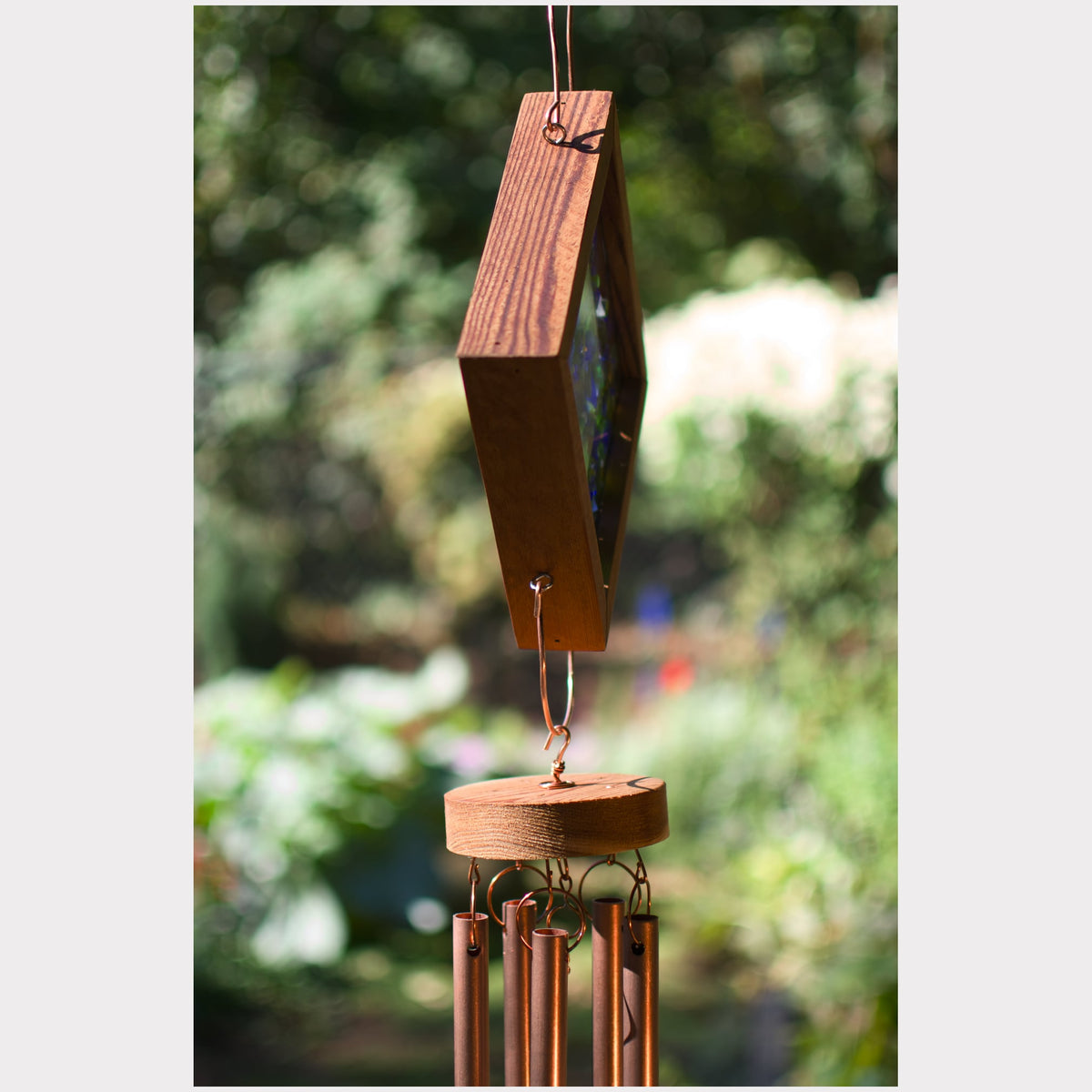 A wooden wind chime hanging outdoors against a blurred green garden background. The chime features a dark brown wood frame with a colorful stained glass panel, a round wooden striker, and several copper-colored metal tubes below.