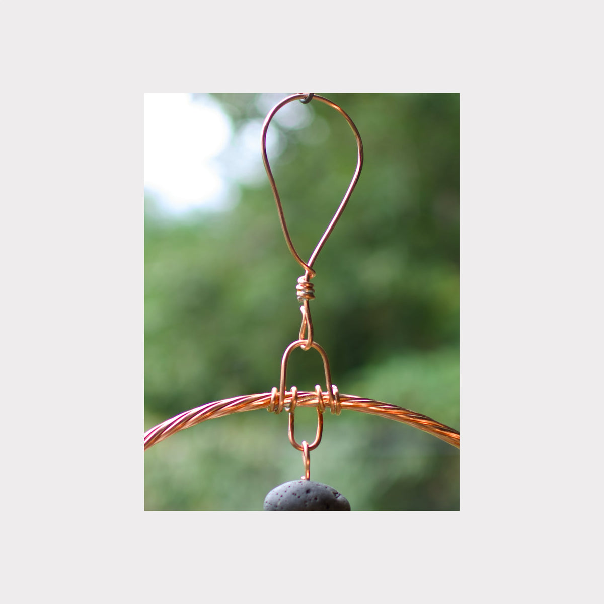 A close-up of a copper wire wind chime with a dark gray stone pendant, suspended against a blurred green outdoor background.