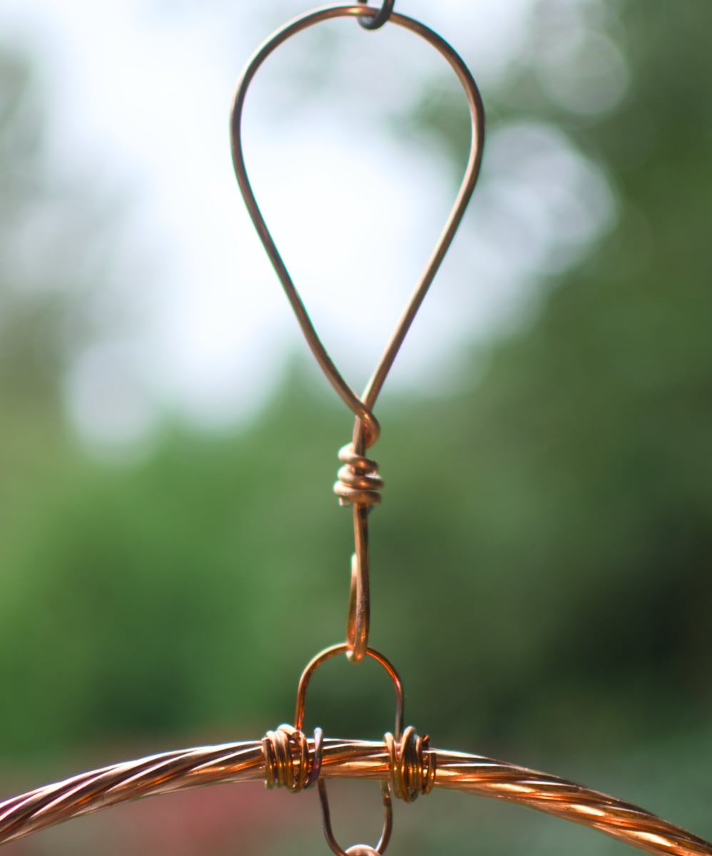 Close-up of a copper-colored metal hanging loop with a twisted wire connector and a thin coiled wire element, set against a blurred green outdoor background.