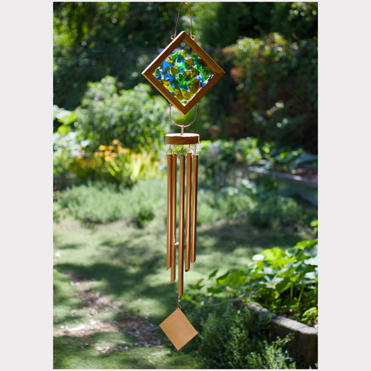 A hanging wind chime with a diamond-shaped stained-glass top, tan metal tubes, and a small square charm, photographed outdoors in a garden setting.