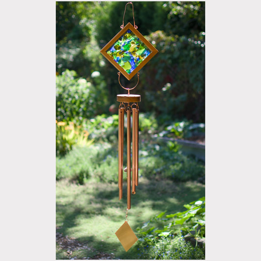 A hanging wind chime with a diamond-shaped stained glass top featuring green, blue, and yellow floral patterns, surrounded by a copper-toned frame. The chime has multiple copper-colored metal tubes and a gold-colored striker, photographed outdoors against a blurred garden background.