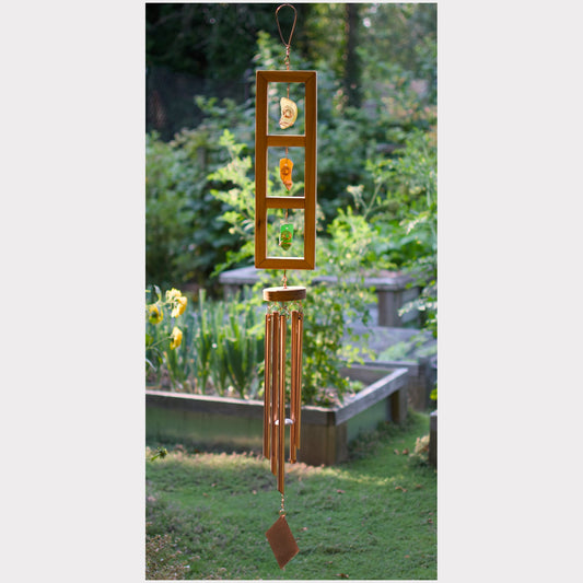 A wooden garden wind chime hanging outdoors in a grassy garden with blurred plants and flowers in the background. The chime features a vertical wooden frame with three rectangular panels containing small colorful decorative elements, three hanging metallic tubes, and a dangling diamond-shaped ornament.