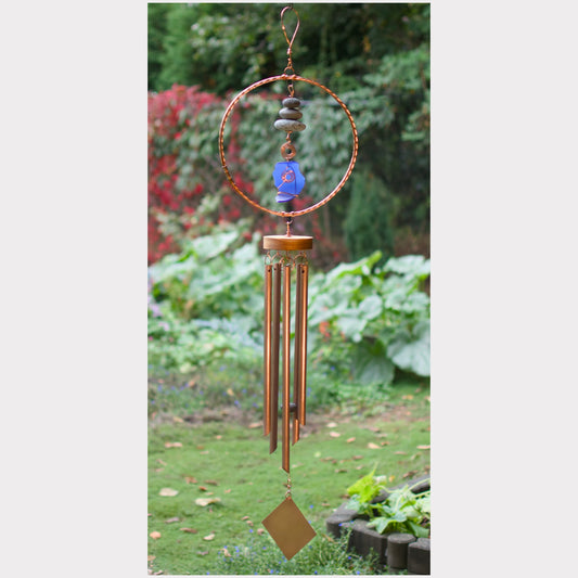 A handmade copper wind chime hanging outdoors in a garden. The wind chime features a circular copper hoop with a blue glass bead, small stones, and a diamond-shaped copper pendant, with several long copper tubes hanging below.