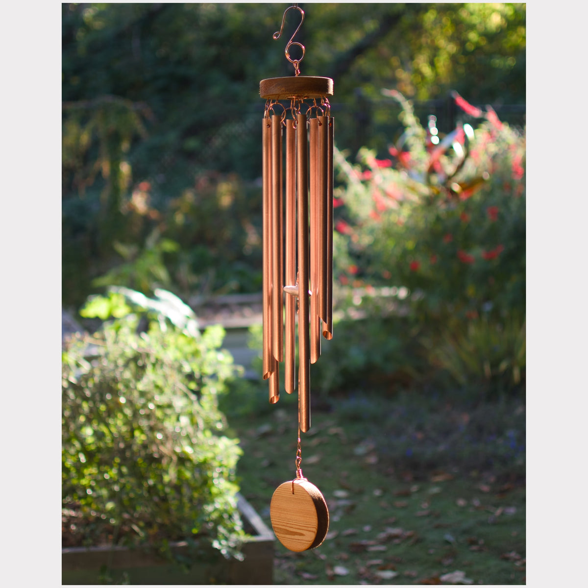 A copper wind chime with multiple hanging tubes and a wooden striker, suspended outdoors against a blurred garden background.