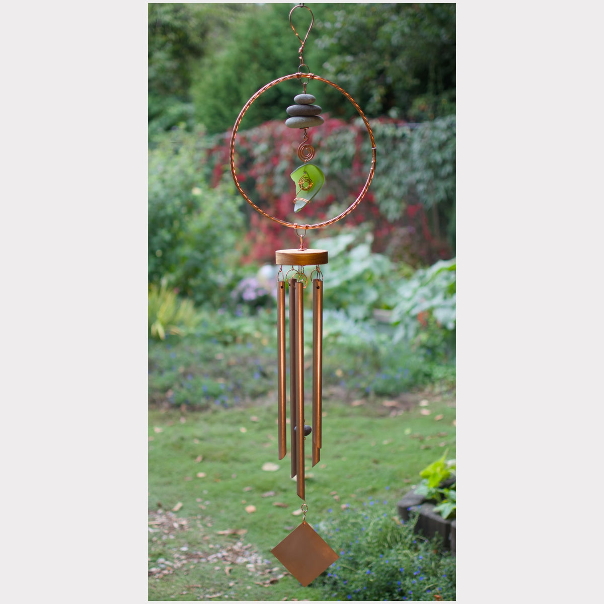 A copper wind chime with stacked stones and a green leaf accent, hanging outdoors against a blurred garden background.