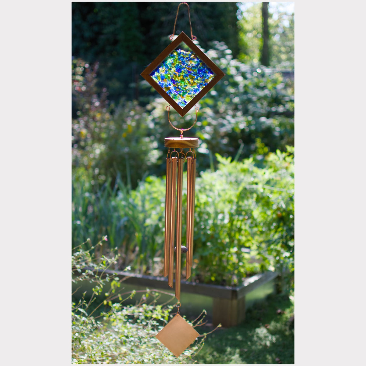 A copper wind chime garden ornament hanging outdoors in a garden. The top section features a diamond-shaped glass panel with blue and green mosaic artwork, suspended from a copper frame, with multiple copper wind chime tubes hanging below and a small diamond-shaped copper pendant at the bottom.
