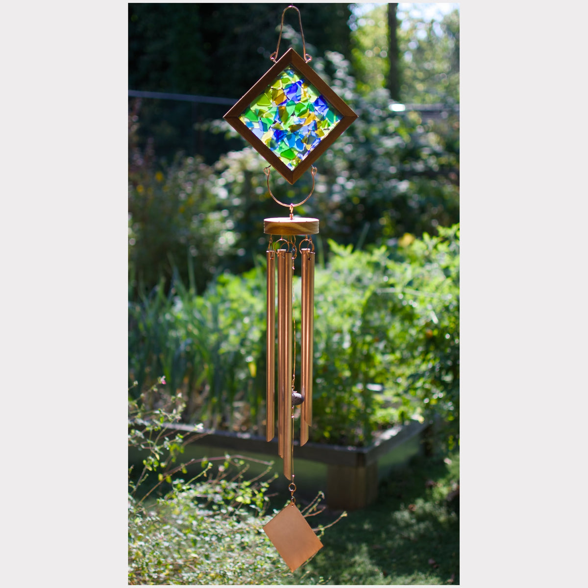 A hanging wind chime with a colorful stained-glass-style diamond top and copper-toned metal tubes, photographed outdoors in a garden setting.