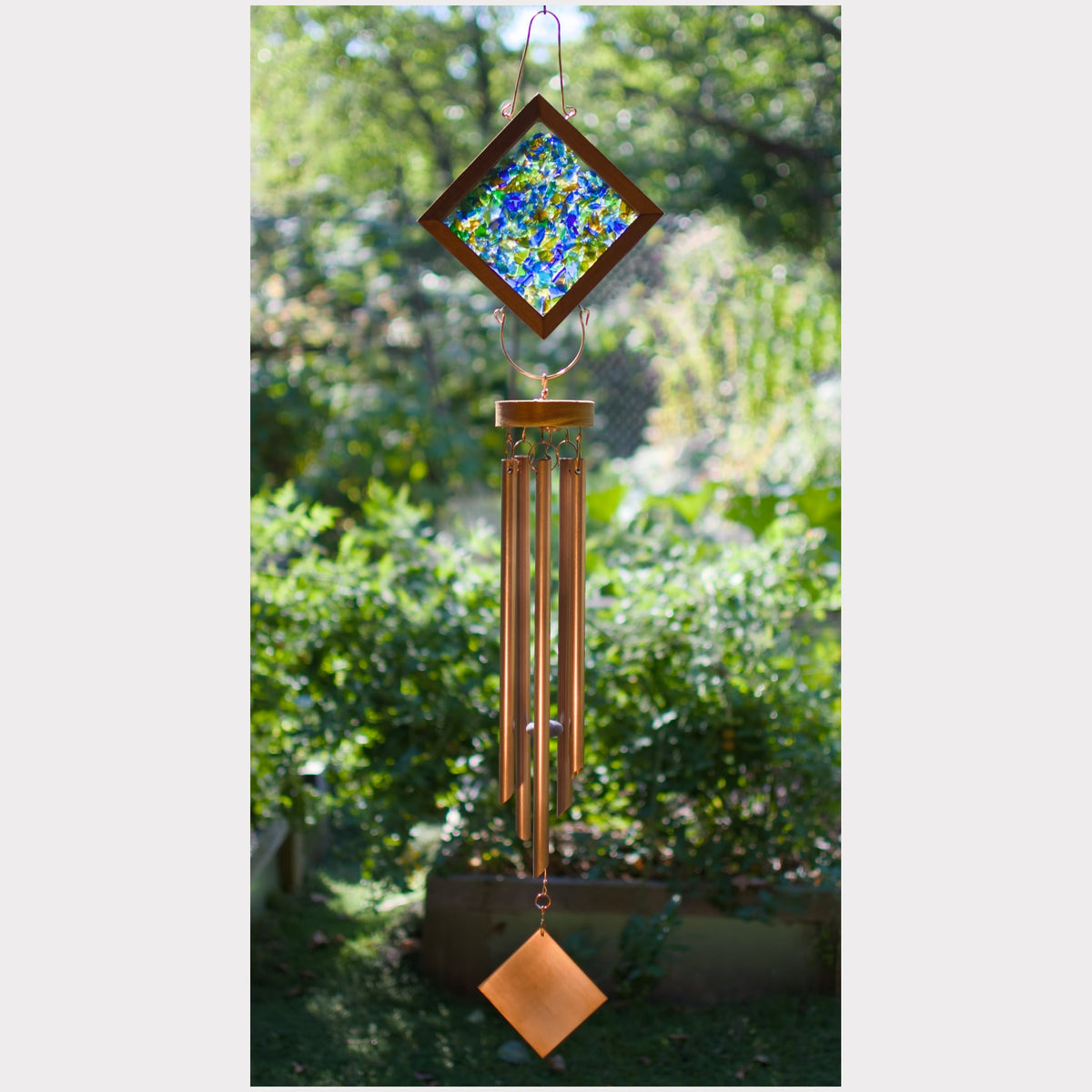 A hanging wind chime with a diamond-shaped stained glass top featuring blue, green, and yellow patterns, copper-toned metal tubes, and a matching diamond-shaped ornament at the bottom, photographed outdoors against a blurred garden background.