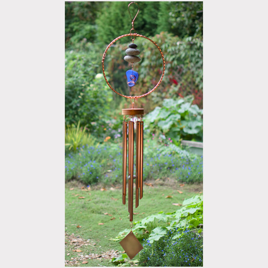 A copper wind chime hanging outdoors in a garden, featuring a circular hoop with stacked stones, a blue glass bead, and multiple copper-colored metal tubes with a diamond-shaped ornament at the bottom.