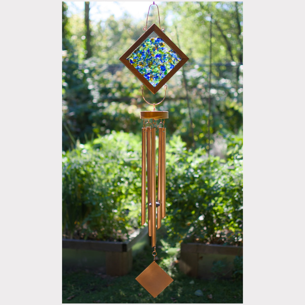 A hanging wind chime with a diamond-shaped stained-glass top featuring blue and green floral patterns, copper-colored metal tubes, and a small diamond-shaped ornament at the bottom, photographed outdoors against a blurred garden background.