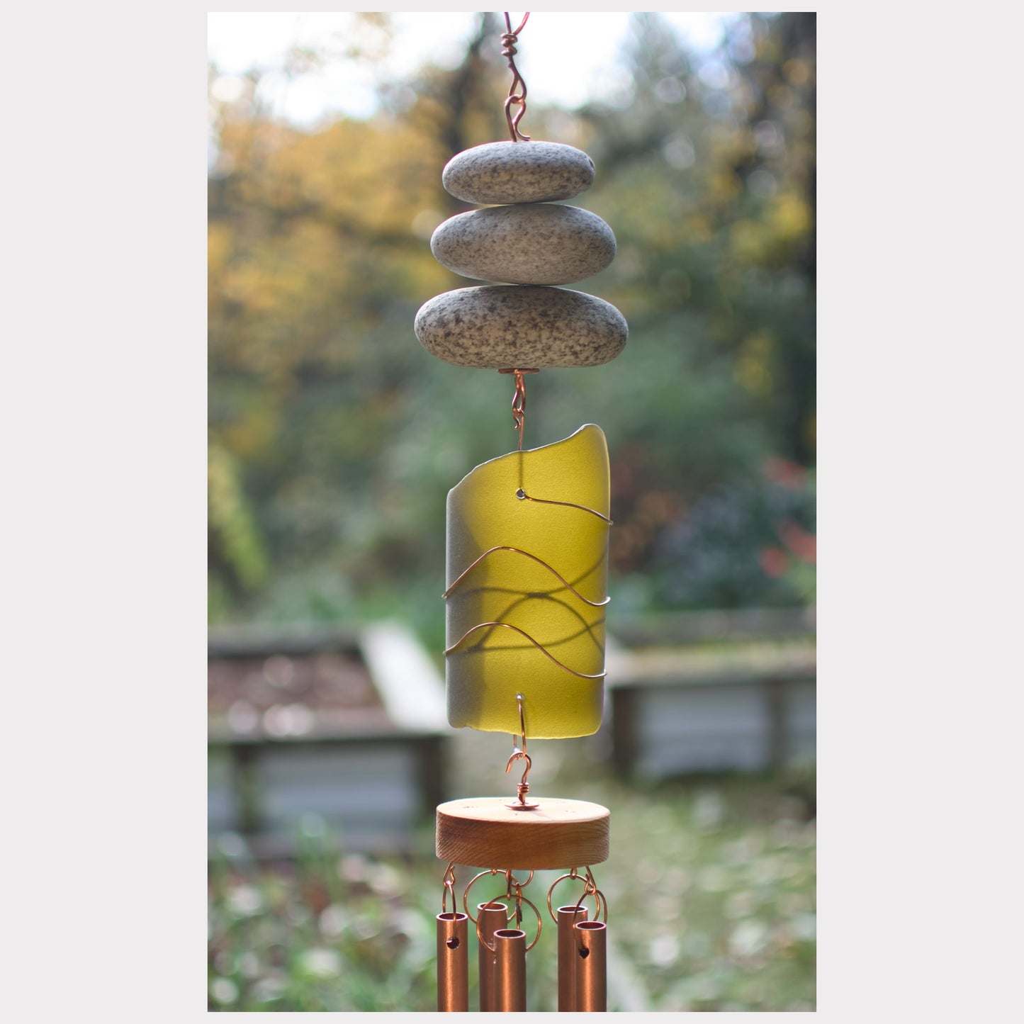 Decorative wind chime with stones and a yellow cylindrical component against a blurred natural background