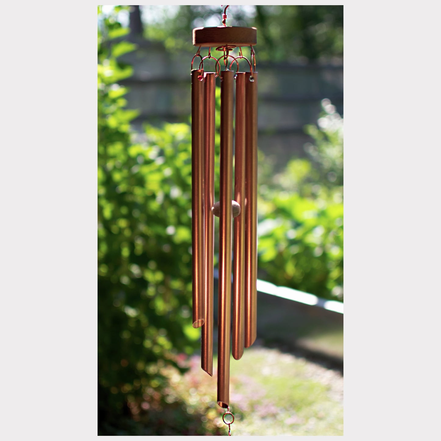 A copper wind chime with multiple hanging tubes and a wooden top cap, photographed outdoors against a blurred green garden background.
