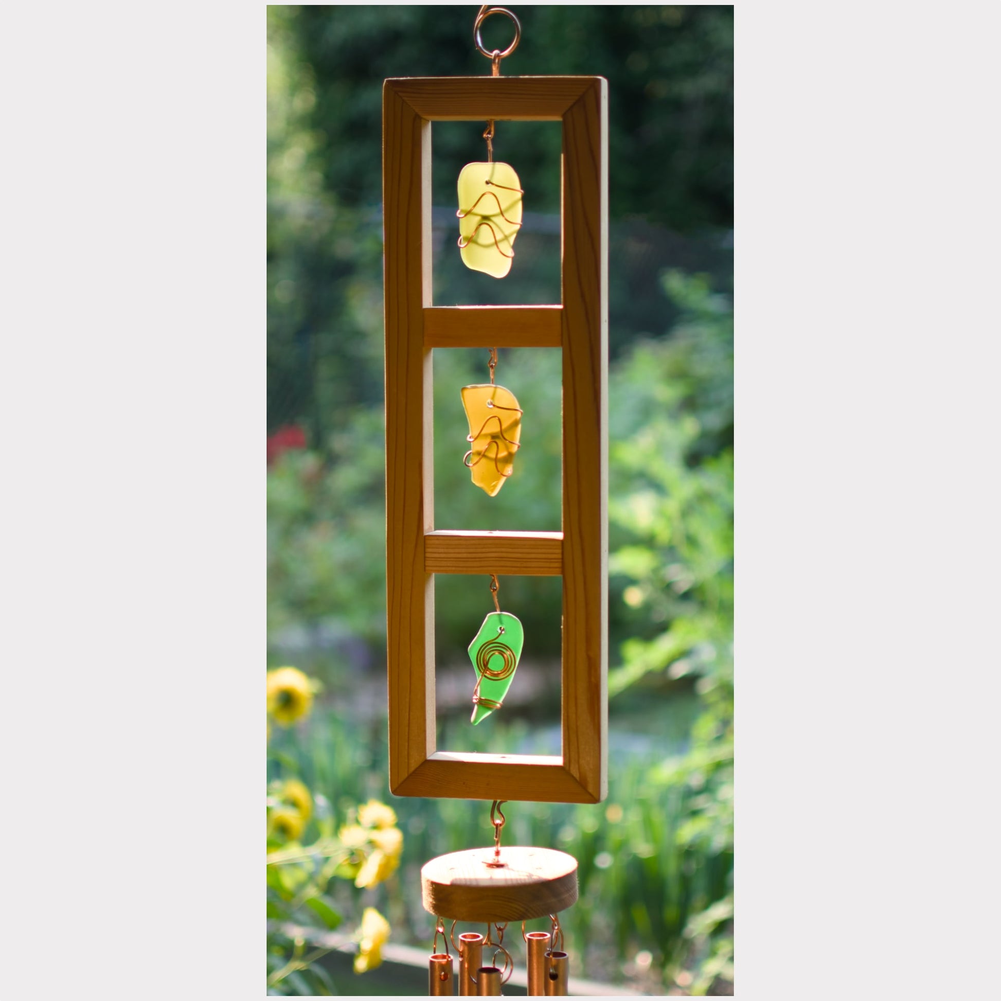 A wooden vertical garden wind chime with three hanging glass panels containing yellow and green leaves, suspended over a round wooden wind chime base against a blurred green garden background.