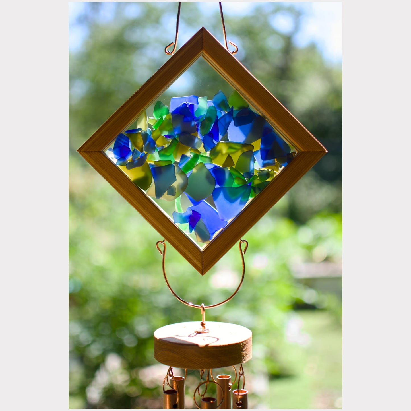 A diamond-shaped wooden wind chime hanging outdoors, featuring a colorful mosaic of blue, green, and yellow glass pieces inside a wooden frame, with copper-colored metal wind chimes below on a blurred green garden background.