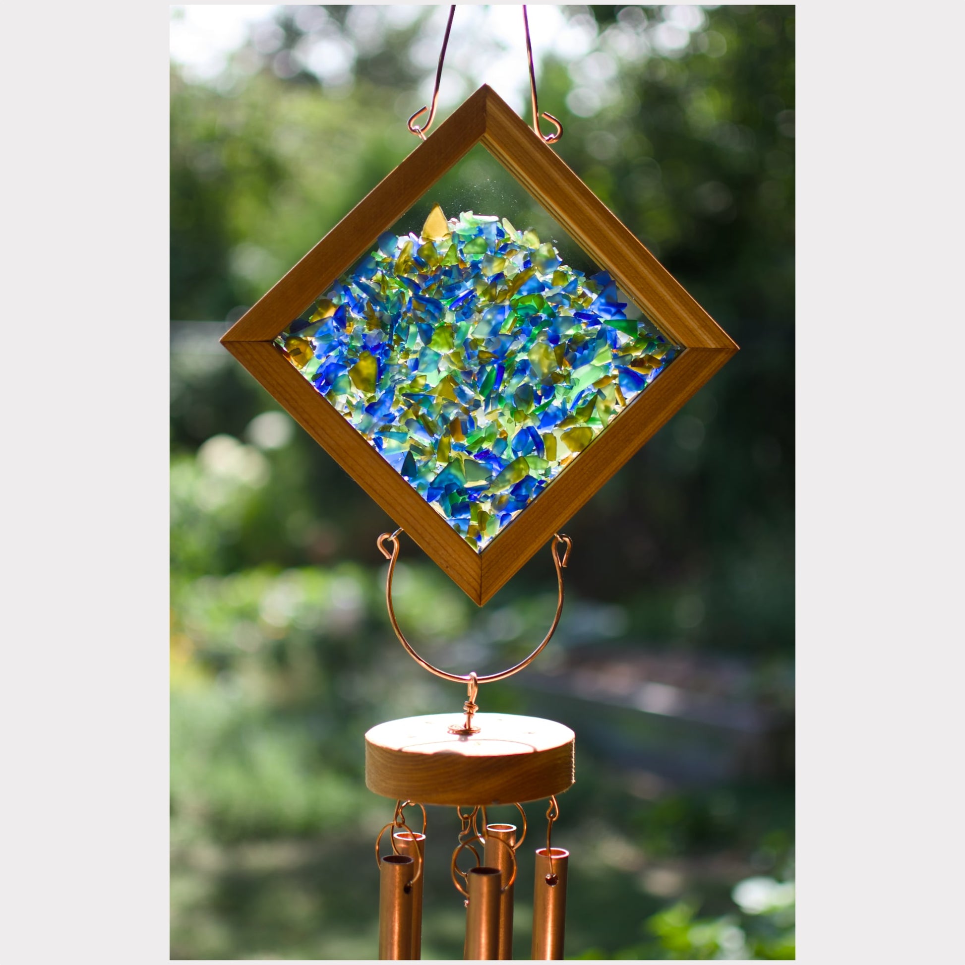 A diamond-shaped wind chime with a wooden frame and colorful blue, green, and yellow glass pieces, hanging outdoors against a blurred green background.