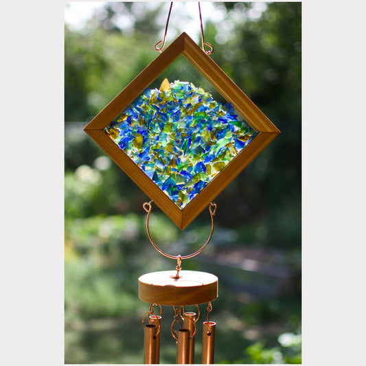 A diamond-shaped wind chime with a wooden frame and colorful blue, green, and yellow glass pieces, hanging outdoors against a blurred green background.