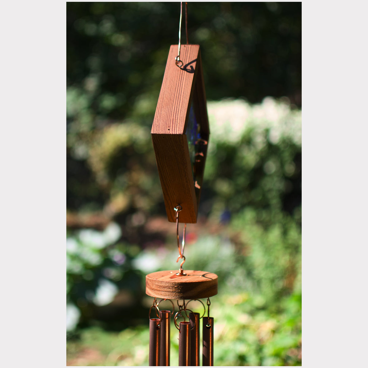 A wooden wind chime hanging outdoors against a blurred green garden background. The chime features a hexagonal wooden top block, a circular wooden striker, and several hanging metallic tubes.