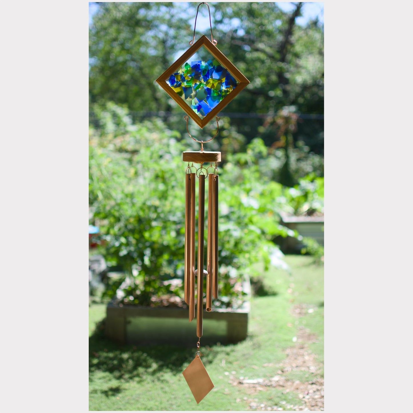 A hanging wind chime with a diamond-shaped stained glass top featuring blue and yellow flowers, four bronze-colored metal tubes, and a small hanging tag, photographed outdoors against a blurred garden background.