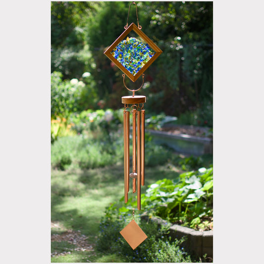 A diamond-shaped wind chime hanging in a garden, featuring a blue and green mosaic pattern in the center, copper-colored metal tubes, and a small diamond-shaped ornament at the bottom.