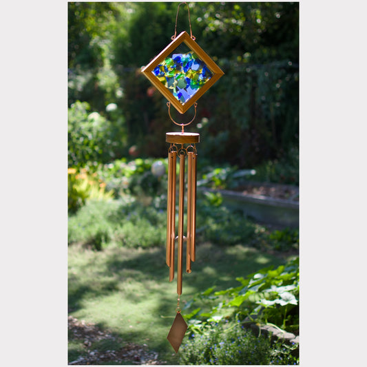 A hanging wind chime with a diamond-shaped stained-glass top featuring blue, green, and yellow floral artwork, surrounded by a gold-toned frame. The chime has multiple copper-colored metal tubes and a dangling ornament, photographed outdoors against a blurred garden background.