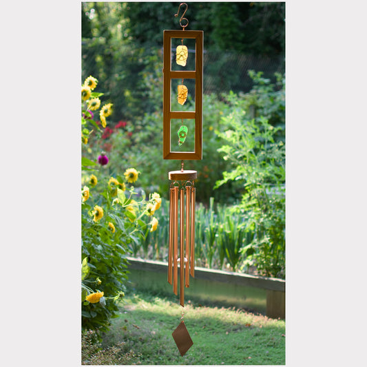 A hanging garden wind chime with a rectangular wooden frame holding three colorful glass panes, suspended above multiple copper-toned metal tubes in an outdoor garden setting.